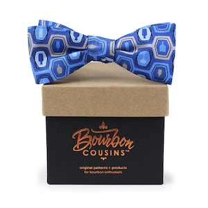 May include: A blue bow tie with a pattern of bourbon bottles in a geometric design. The bow tie is in a brown box with the text "Bourbon Cousins" in gold lettering.