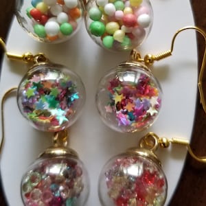 May include: Six pairs of dangle earrings with clear, spherical charms. The charms are filled with colorful beads and star-shaped confetti. The earrings have gold-colored hooks and accents, displayed on a white oval card.