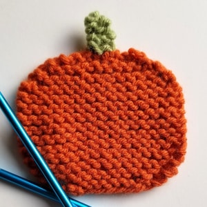 May include: A knitted orange pumpkin with a green stem, resting on a white surface with two blue knitting needles.