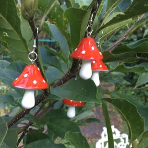 May include: Red and white mushroom earrings with a polka dot design, hanging from a tree branch. The earrings have a bright red cap with white spots and a white stem. The background features green leaves and a blurred outdoor setting.
