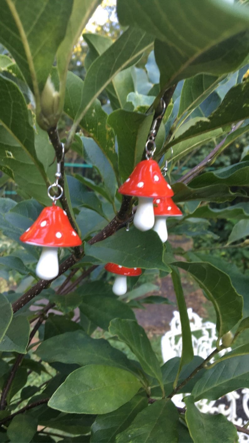 May include: Red and white mushroom earrings with a polka dot design, hanging from a tree branch. The earrings have a bright red cap with white spots and a white stem. The background features green leaves and a blurred outdoor setting.