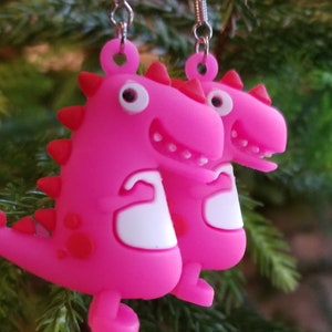 May include: A pair of pink dinosaur earrings with white teeth and white eyes. The dinosaurs are holding their arms up in the air.