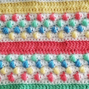 May include: Close-up of a colorful, handmade crochet blanket. The blanket features rows of white, yellow, pink, blue, and green yarn. The design includes textured bobbles and stripes, creating a visually appealing and cozy textile.