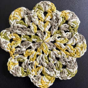 May include: A circular crochet doily featuring a floral-inspired pattern with six rounded petals. The piece is crafted from variegated yarn in shades of mustard yellow, olive green, cream, and light brown, creating a textured, openwork design.