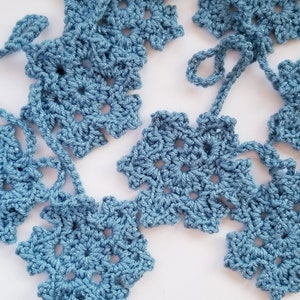 May include: A garland of crocheted snowflakes in a light blue color. The snowflakes are connected by a string of yarn.