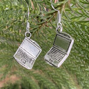 May include: A pair of silver-colored laptop earrings. Each earring features a detailed miniature laptop design with a screen, keyboard, and trackpad. The earrings hang from silver-colored fish hook ear wires.