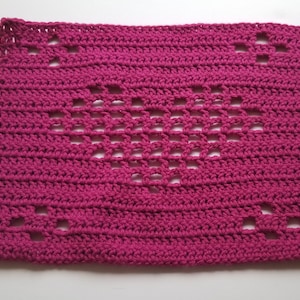 May include: A pink crocheted square with a heart-shaped design in the centre. The heart is created by a series of open stitches.
