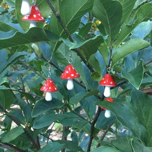 May include: Red and white mushroom earrings hanging from silver hooks. The earrings are set against a backdrop of green leaves and branches. The mushroom caps are red with white spots, and the stems are white.