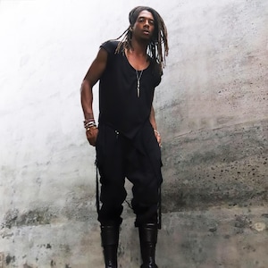 May include: A person wearing a black outfit, including a short-sleeved top, pants, and knee-high platform boots. The outfit is accessorized with a necklace and bracelets. The background is a textured gray wall.