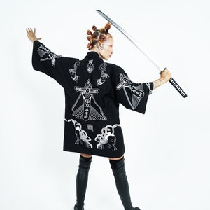 May include: A person wearing a black kimono with white embroidered designs, holding a silver katana sword. The kimono features a geometric pattern and a stylized image of a person's face.