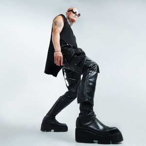 K&ouml;nnte beinhalten: Eine Person tr&auml;gt ein schwarzes Tanktop, eine schwarze Lederhose und schwarze Stiefel. Die Person hat einen rasierten Kopf und tr&auml;gt eine Sonnenbrille.