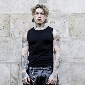 K&ouml;nnte beinhalten: Ein Mann mit Tattoos und gedehnten Ohrl&auml;ppchen tr&auml;gt ein schwarzes &auml;rmelloses Shirt und eine schwarze Lederhose mit Cargo-Taschen.