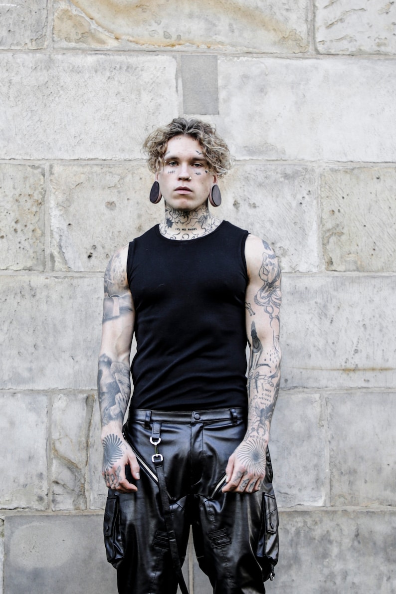 K&ouml;nnte beinhalten: Ein Mann mit Tattoos und gedehnten Ohrl&auml;ppchen tr&auml;gt ein schwarzes &auml;rmelloses Shirt und eine schwarze Lederhose mit Cargo-Taschen.