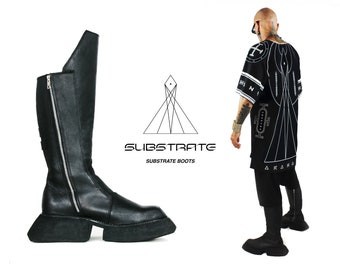 Op de afbeelding: Zwarte leren laarzen die tot aan de knie komen met een dikke platformzool en een zijrits. De laarzen staan op een witte achtergrond met de tekst "SUBSTRATE SUBSTRATE BOOTS" en een geometrisch ontwerp.