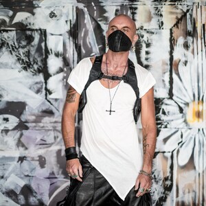May include: A man wearing a white t-shirt and black leather pants stands in front of a graffiti wall. He is wearing a black leather face mask with a silver nose ring and a silver chain necklace with a cross pendant.