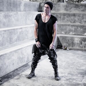 May include: A man wearing a black beanie, black t-shirt, black pants, and black boots. He is standing in front of a concrete wall with steps.