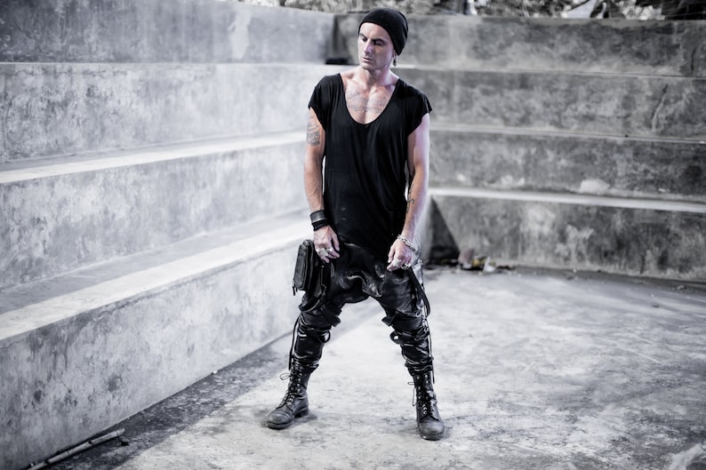 May include: A man wearing a black beanie, black t-shirt, black pants, and black boots. He is standing in front of a concrete wall with steps.