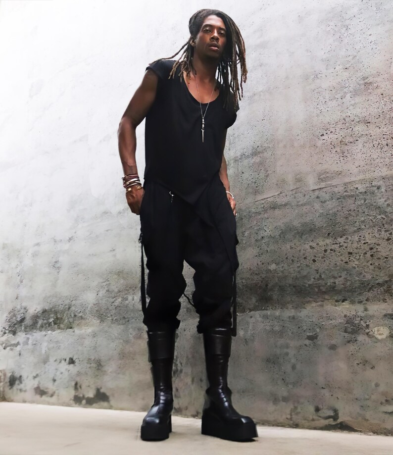 May include: A person wearing a black sleeveless shirt, black pants with straps, and black boots. The person has long dreadlocks and is standing in front of a concrete wall.