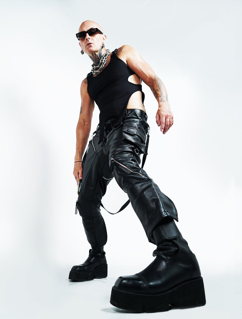 K&ouml;nnte beinhalten: Ein kahlk&ouml;pfiger Mann mit schwarzer Sonnenbrille, einem schwarzen Tanktop, einer schwarzen Lederhose und schwarzen Plateauschuhen. Er tr&auml;gt eine silberne Kettenkette und Tattoos auf seinen Armen.
