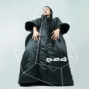 May include: A person wearing a black puffer jacket with a long, flowing design. The jacket has a high collar and a zipper closure. The person is standing in front of a white background with a geometric shape drawn around them. The text "e2ad" is visible in the image.