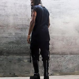 May include: A person wearing a black sleeveless shirt and black pants with long straps. The person is wearing black boots with a zipper on the back.