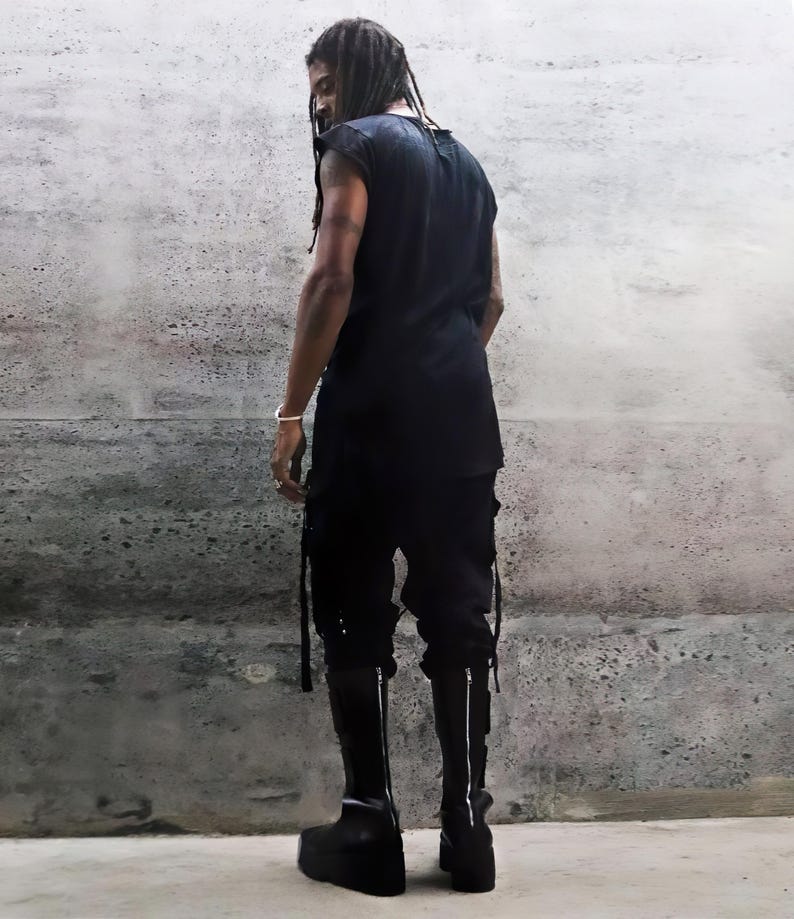 May include: A person wearing a black sleeveless shirt and black pants with long straps. The person is wearing black boots with a zipper on the back.