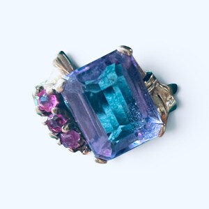 May include: A ring featuring a large, rectangular, emerald-cut stone with shifting blue and purple hues. The central stone is flanked by three small, round, deep pink stones on one side, all set in a metallic band with a textured, leaf-like design.