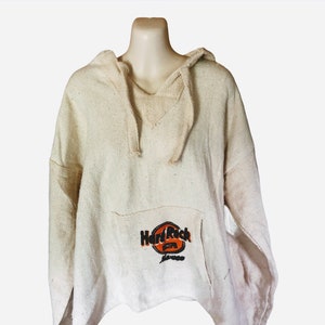 May include: Off-white, hooded pullover with a front pocket. The pocket features the Hard Rock Cafe logo in orange, black, and white. The top has a V-neck and drawstring hood. The material appears to be a woven fabric.