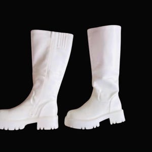 May include: A pair of white, knee-high boots with a chunky sole. The boots have a smooth, matte finish and a ridged detail on the upper shaft. The boots are set against a black background.