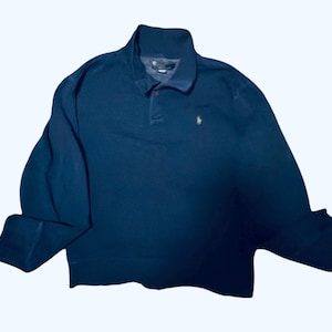 May include: A navy blue long-sleeved polo shirt with a button-down collar. The shirt features a small, gold-colored logo on the chest. The sleeves are cuffed, and the shirt is made of a soft material.