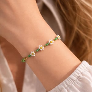 May include: A beaded bracelet featuring a daisy flower design. The bracelet has white and yellow daisy flowers, green beads, and gold-colored accents. The bracelet is worn on a wrist.