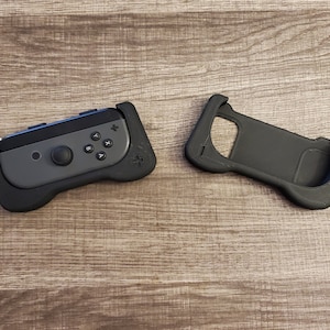 May include: Black and gray gaming controller grip with a black wrist strap. The controller has a gray face with black buttons and a joystick. A separate black grip is also shown.