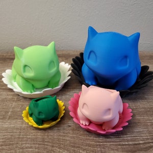 May include: Four Bulbasaur planters in various colors: green, blue, pink, and dark green. Each planter is sitting in a flower-shaped base. The planters are arranged on a wooden surface.