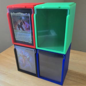 Peut inclure: Quatre boîtes de rangement pour cartes en rouge, vert, bleu et noir. Chaque boîte a une façade en plastique transparent et est conçue pour contenir des cartes à collectionner. Les boîtes sont empilées les unes sur les autres.
