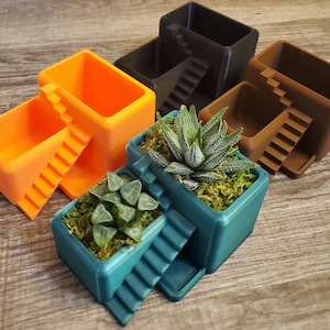 May include: A selection of 3D-printed plant pots in shades of orange, teal, brown, and black. Each pot has a staircase design. Some pots are filled with small succulents and moss.