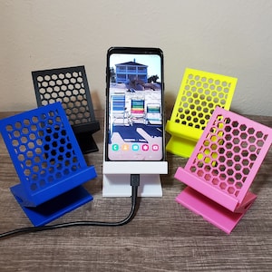 May include: A smartphone is displayed on a white phone stand with a charging cable. Surrounding the phone are four colorful phone stands in blue, black, yellow, and pink. Each stand has a honeycomb pattern.