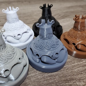 May include: Five 3D-printed decorative figurines in white, black, grey, brown, and a lighter grey. Each figurine features an ornate design, a rounded base, and a crown-like top. The figurines are displayed on a wooden surface.