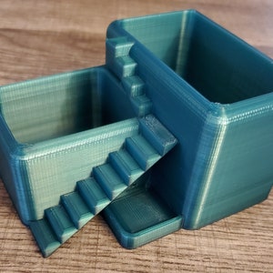 May include: A teal-colored, 3D-printed desk organizer with two rectangular compartments connected by a staircase. The organizer is made of plastic and has a textured surface. The design is modern and geometric.