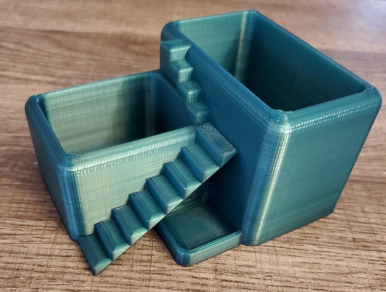 May include: A teal-colored, 3D-printed desk organizer with two rectangular compartments connected by a staircase. The organizer is made of plastic and has a textured surface. The design is modern and geometric.