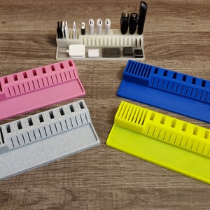 May include: A collection of 3D-printed desk organizers in pink, gray, blue, and yellow. Each organizer features a slotted design for holding various items, with a separate section for smaller objects. A larger organizer holds various tech accessories.