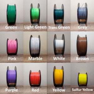 May include: A collection of 15 spools of 3D printer filament in various colors, including black, green, pink, and blue. Each spool is labeled with its color name. The spools are arranged in rows on a wooden surface.