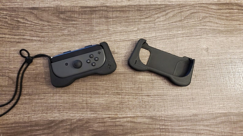 May include: Black and gray gaming controller grip with a black wrist strap. The controller has a gray face with black buttons and a joystick. A separate black grip is also shown.