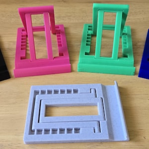 May include: An assortment of colorful phone stands in black, pink, green, and blue. The stands are made of plastic and have an adjustable design. A light purple stand is also included.