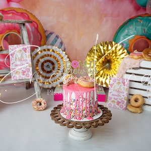 May include: A pink and white donut-themed cake with sprinkles and pink frosting. The cake is decorated with a gold glitter topper that says "Donut Grow Up". The cake is on a wooden cake stand and surrounded by donuts, paper fans, and a wooden ladder.