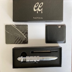 May include: A silver tactical pen with a black case and a black leather wallet. The pen has a silver clip and a black handle. The wallet has a silver clip and a black leather exterior. The pen and wallet are both labeled with the text "GG Tactical".