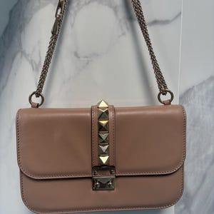 Op de afbeelding: Blushkleurige leren schoudertas met een kettingriem. De klepsluiting is versierd met goudkleurige piramidevormige studs. De tas heeft een rechthoekige vorm en een zilverkleurige sluiting.