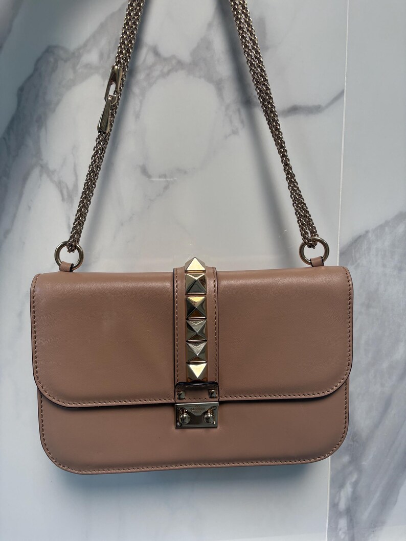 Op de afbeelding: Blushkleurige leren schoudertas met een kettingriem. De klepsluiting is versierd met goudkleurige piramidevormige studs. De tas heeft een rechthoekige vorm en een zilverkleurige sluiting.