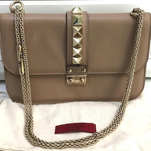 Op de afbeelding: Een beige leren schoudertas met goudkleurige kettingriemen. De klepsluiting heeft een rij gouden piramide studs. De tas heeft een gouden sluiting en een klein rood label met de merknaam.