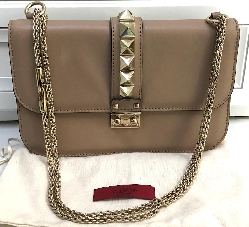 Op de afbeelding: Een beige leren schoudertas met goudkleurige kettingriemen. De klepsluiting heeft een rij gouden piramide studs. De tas heeft een gouden sluiting en een klein rood label met de merknaam.