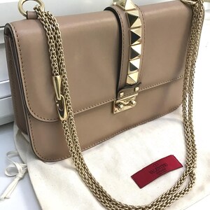Op de afbeelding: Beige leren handtas met goudkleurige kettingriemen en piramidevormige studs. De klepsluiting heeft een gouden sluiting. Een rood label met witte tekst is zichtbaar.